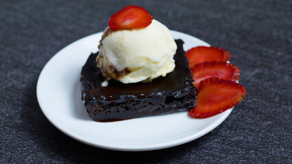 Chocolate Brownie Sundae with chocolate sauce, vanilla ice cream scoop and fresh sliced strawberries are served on a white plate, creating a tempting dessert
