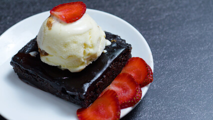 Chocolate Brownie Sundae with chocolate sauce, vanilla ice cream scoop and sliced strawberries, served on a white plate over a dark gray table, representing a sweet and delicious dessert