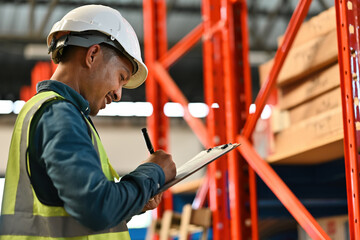 A male warehouse worker in safety gear checks and records inventory data on a clipboard, representing accuracy, stock control, and industrial efficiency in a professional storage environment