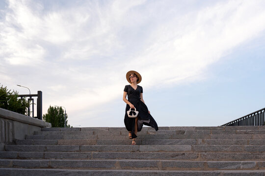 Stylish woman descends stone stairs holding handbag and hat