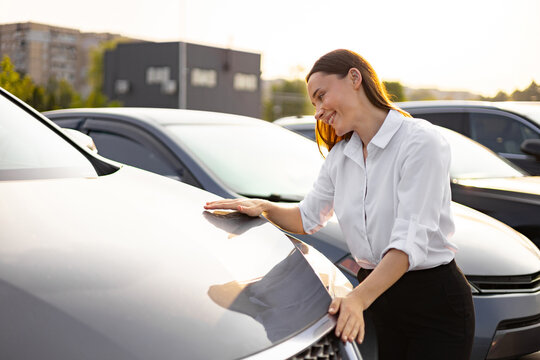 A smiling woman is looking at a new car, touching its hood, and considering a purchase at a dealership. - Powered by Adobe