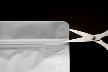 A pair of silver scissors positioned to cut open the top of a white, blank packaging pouch along its dashed tear line, against a dark background