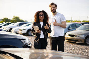 A female car dealer and a male customer smile and give a thumbs up in front of a car dealership.
