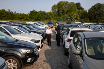 A male car dealer in a suit showing a car to a woman at a dealership, with various cars in the background. © Sofiia Shunkina