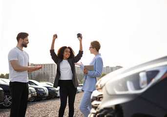 A happy Black woman celebrates buying a new car, raising keys and a coffee cup, with a car salesperson and a man in the background.