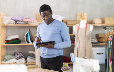Thoughtful African American male dressmaker working on digital sketch of new garment in workshop surrounded by fabric, mannequins, and sewing equipment
