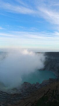 Aerial view of steaming turquoise acidic crater lake at Kawah Ijen volcano in East Java, Indonesia, sulfur haze rising at sunrise over landscape.