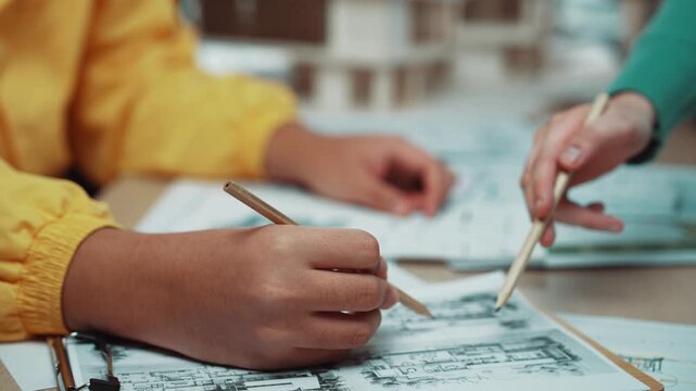A designer in a vibrant yellow jacket is focusing on sketching architectural plans with a pencil. The workspace is filled with various tools and documents, enhancing creativity. SACTR