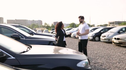 A female car dealer hands car keys to a male customer in a car dealership lot under sunlight.