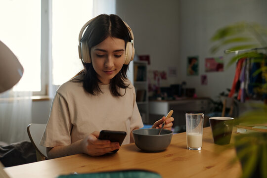 Caucasian teenage girl sitting at table listening to music with headphones, while holding smartphone and eating breakfast cereal with glass of milk in front of her