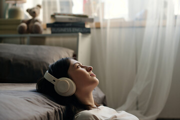 Caucasian young adult woman lying on floor listening to music with headphones, gazing upward near...