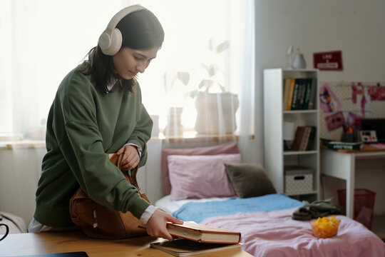 Teenage Caucasian girl wearing headphones packing books into backpack in bedroom, standing near desk with bed and shelves in background, preparing for school or study session