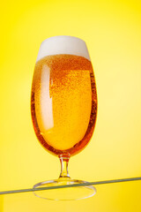 A tall glass of beer with a golden hue and frothy head, on a yellow background