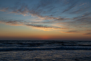 Fototapeta premium Dramatic sunset over a stormy sea with dark clouds, golden-orange horizon, and waves crashing onto the sandy shore, reflecting the warm colors of the sky.Keywords: