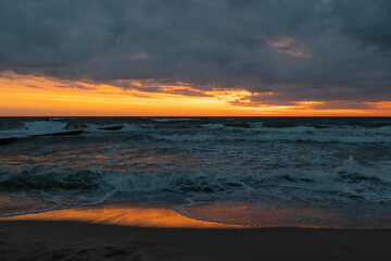 Fototapeta premium Dramatic sunset over a stormy sea with dark clouds, golden-orange horizon, and waves crashing onto the sandy shore, reflecting the warm colors of the sky.