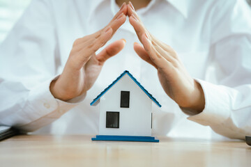 Hands carefully hold and shield a small house model, symbolizing protection, home insurance,...