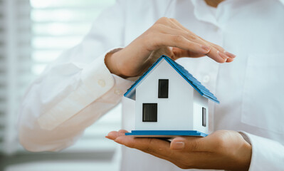 Hands carefully hold and shield a small house model, symbolizing protection, home insurance,...