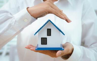 Hands carefully hold and shield a small house model, symbolizing protection, home insurance,...