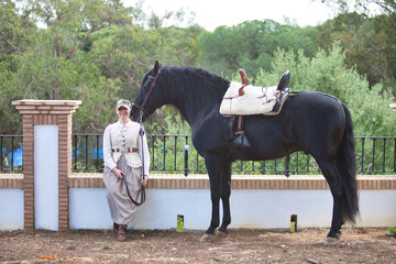 Beautiful young woman sitting in the shade of a pine tree next to her black purebred Spanish horse...
