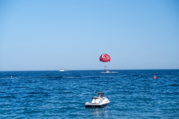 Bright sunny day features a person parasailing high above the ocean while a jet ski floats nearby,...