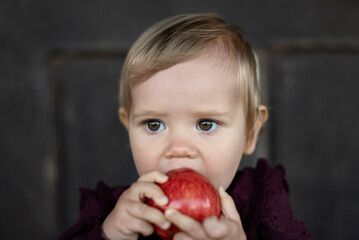 A close-up of a toddler eating a red apple, in front of wooden house.