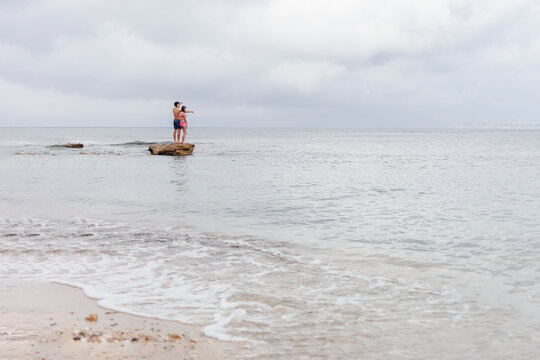 Two teens stand on rock at edge of sea gazing at ocea