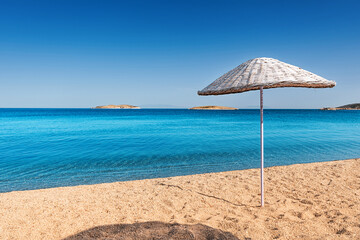 Straw beach umbrella standing on a deserted beach with turquoise water and small islands in the background under a clear blue sky
