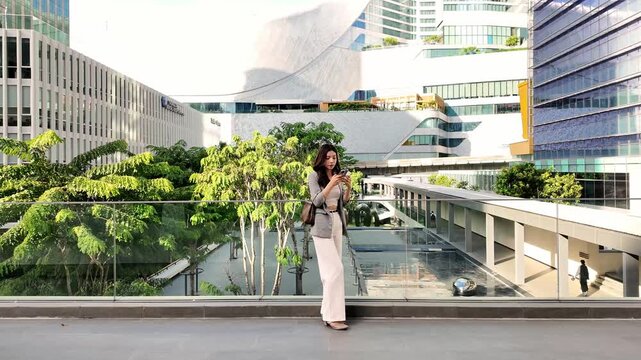 Elegant young woman in business attire stands on a modern skywalk in a city, holding coffee and using her phone. Urban architecture and greenery frame a blend of nature and contemporary lifestyle.