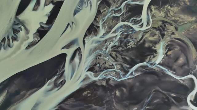 Dynamic aerial view of pale blue and white glacial rivers weaving through dark volcanic sands in Iceland, showcasing intricate natural patterns.