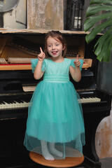 Little happy funny girl in blue dress standing on chair near black piano in room