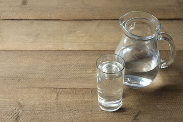 Glass of water with jug at wooden table, closeup. Drinking clear water in the morning for health. Space for text 