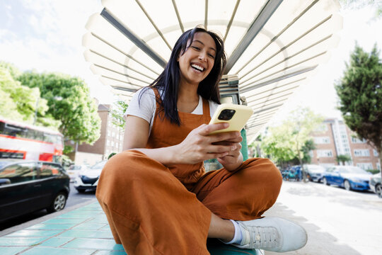 Laughing woman using smartphone at city bus stop outdoors