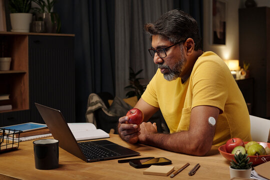 Man at home with insulin patch eating apple while working on laptop