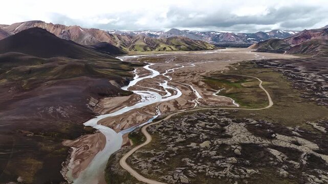 Aerial view of Iceland's Landmannalaugar region with multicolored rhyolite mountains, braided rivers, rocky outcrops, and green patches in motion.