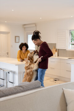 African American couple holding mug and man bending down petting golden retriever by kitchen island