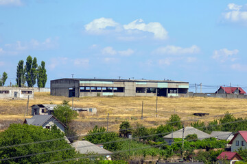 Fototapeta premium Old concrete warehouse in a rural area surrounded by dry grass, trees, and scattered village houses under a blue sky.