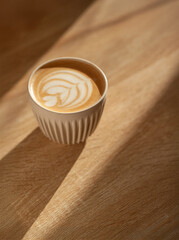 A ceramic cup of cappuccino with elegant cream latte art sits on a light wooden table in a minimalist coffee shop setting. The soft lighting and clean composition highlight the warm tones and inviting