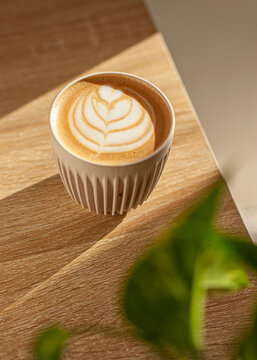 A ceramic cup of cappuccino with elegant cream latte art sits on a light wooden table in a minimalist coffee shop setting. The soft lighting and clean composition highlight the warm tones and inviting