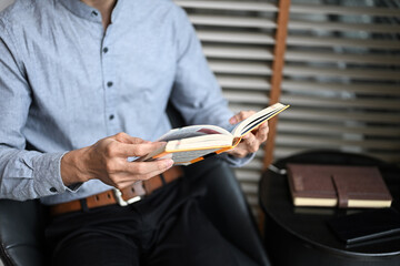 Close up of businessman reading book in cozy chair by the window in a modern office interior