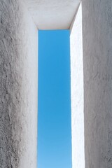 Architectural shot featuring an upward view between light-colored, textured walls revealing a bright blue sky. Minimalist and geometric.