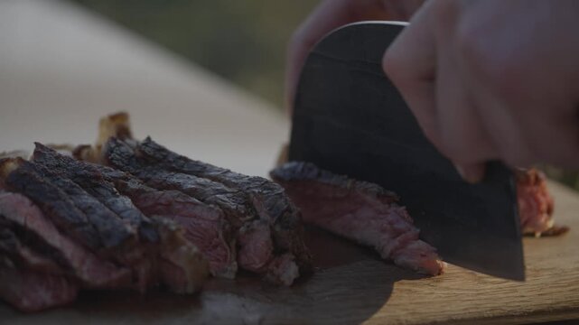 A person is cutting a piece of meat with a knife on a wooden cutting board. The meat is cut into thin slices, and the knife is positioned above the meat. Concept of preparation for a meal