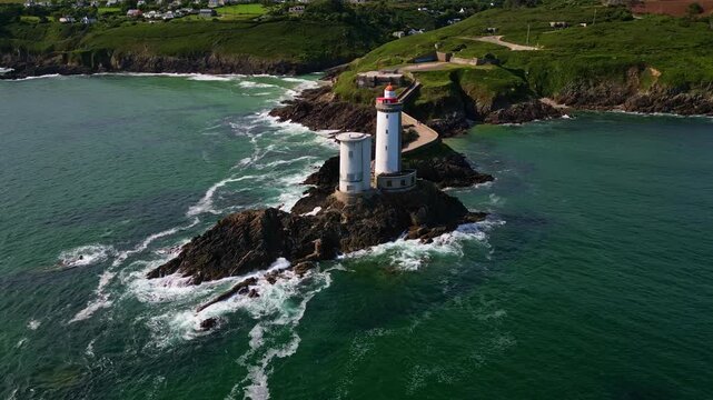 Drone performs a smooth orbit around the Petit Minou lighthouse, showing crashing waves, cliffs, vegetation, and houses under a bright blue sky - Plouzan&eacute;, Brittany in France