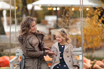 Happy mother and daughter sitting on a swing in an autumn park against the background of pumpkins. Halloween. Motherhood, Mother's Day.