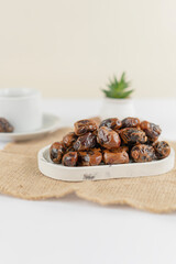 A neat pile of dried dates placed on a marble tray atop a burlap mat, with a cup and a small plant blurred in the background, under soft natural lighting.
