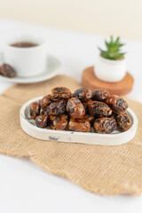 A neat pile of dried dates placed on a marble tray atop a burlap mat, with a cup and a small plant blurred in the background, under soft natural lighting.