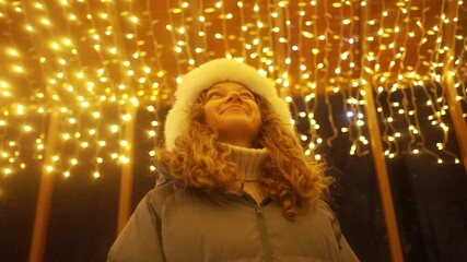 Cheerful young woman standing amid glowing christmas lights. Wearing warm winter clothing. Enjoying magical holiday atmosphere during festive evening street market celebration - Powered by Adobe
