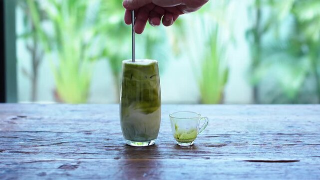 A hand stirs a refreshing iced matcha latte, blending vibrant green tea with cold milk in a tall glass. A small empty cup with matcha residue rests nearby on a rustic wooden table.
