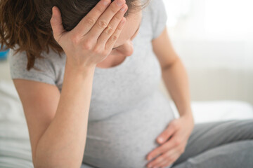 Expecting mother holding her head in pain while resting on bed. Difficult moment of pregnancy migraine and maternal health concern at home.
