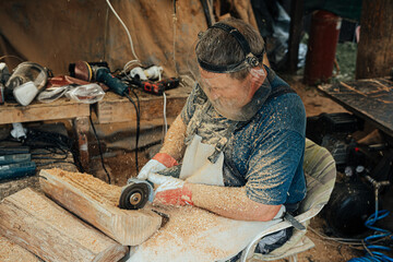 A focused craftsman uses a power tool to carve wood, surrounded by sawdust and tools, wearing full protective gear in a busy workshop