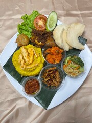 A complete Indonesian yellow rice dish. The plate is decorated with a variety of traditional side dishes, including shredded fried chicken, omelet slices, fried noodles, spicy potatoes, meatballs